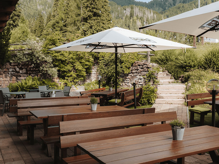 Terrasse im Freien mit Holztischen, weißen Sonnenschirmen und Topfpflanzen. Im Hintergrund sind Bäume und Steinstufen zu sehen. - Hotel Steinerhof