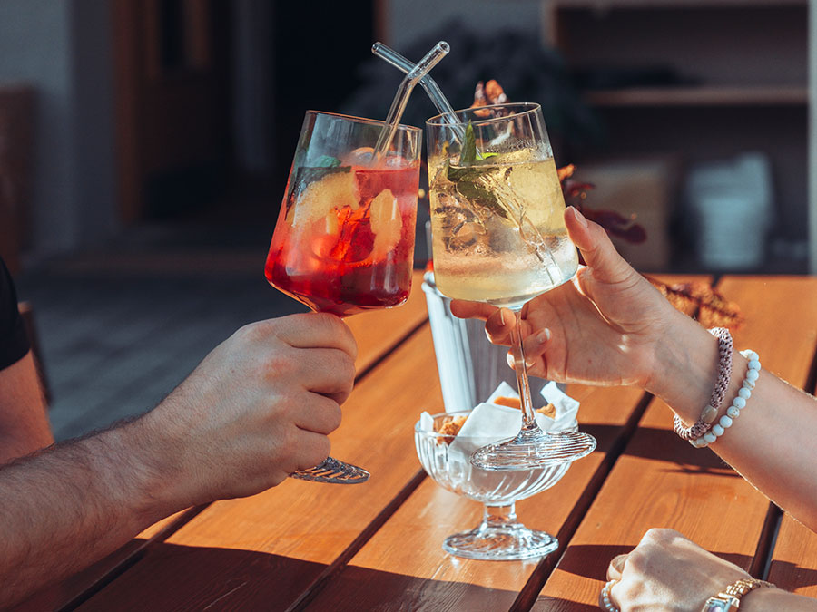 Zwei Personen stoßen mit bunten Cocktails in Gläsern mit Strohhalmen an einem Holztisch im Freien an. - Hotel Steinerhof