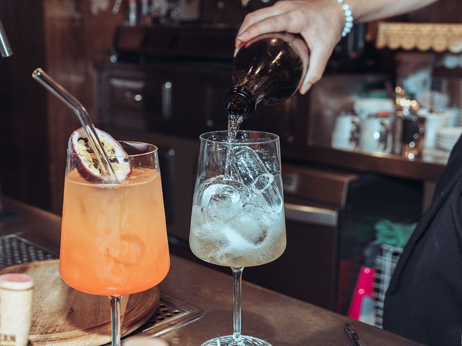 Er gießt das Getränk von Hand in ein Glas mit Eis; daneben steht ein weiterer Cocktail mit Passionsfrucht auf der Theke. - Hotel Steinerhof