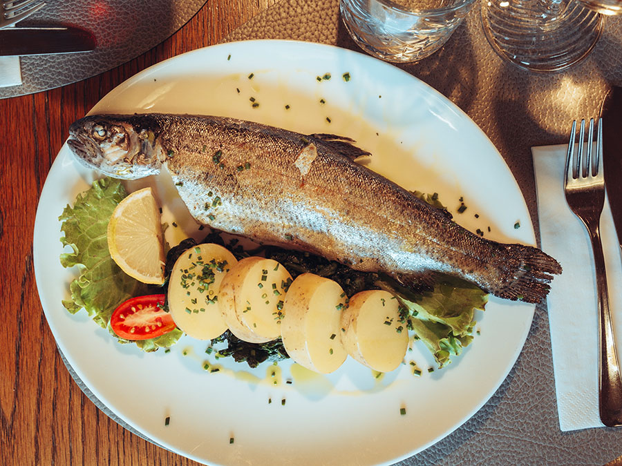 Ein ganzer gegrillter Fisch mit Kartoffelscheiben, Zitrone, Tomate und Blattgemüse auf einem weißen Teller, daneben liegt das Besteck. - Hotel Steinerhof