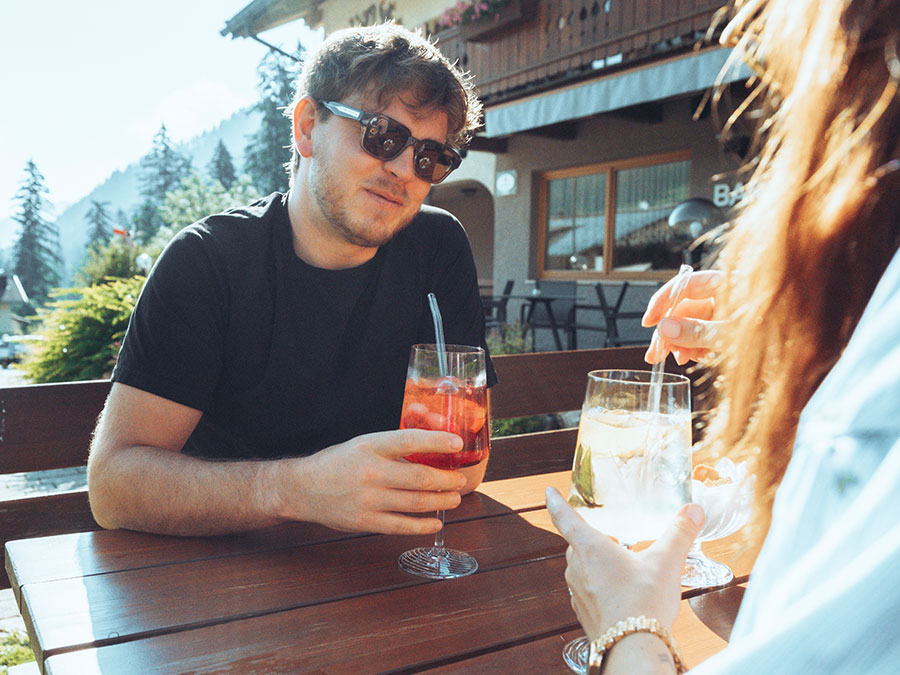 Ein Mann mit Sonnenbrille, der ein Getränk in der Hand hält und an einem sonnigen Tag mit einer anderen Person draußen an einem Holztisch sitzt. - Hotel Steinerhof