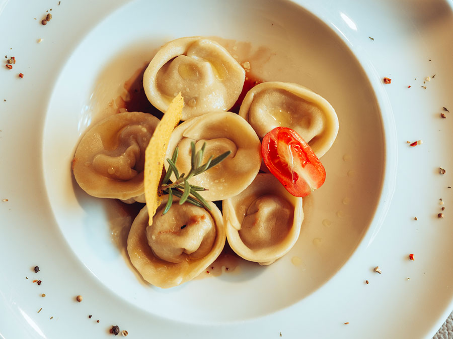 Sechs Teigtaschen, garniert mit einer Tomatenscheibe, Zitronenschale und Rosmarin, auf einem weißen Teller. - Hotel Steinerhof