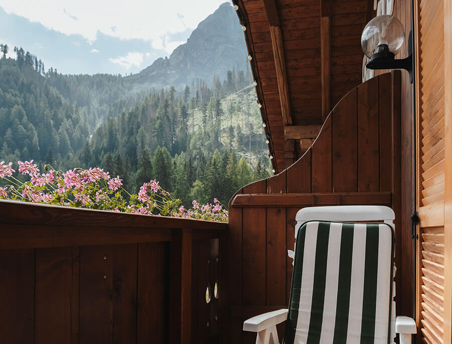 Ein gestreifter Liegestuhl auf einem Holzbalkon mit rosa Blumen und Blick auf einen bewaldeten Berg. - Hotel Steinerhof
