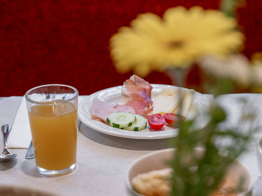 Ein Teller mit Aufschnitt, Käse, Gurke und Tomaten neben einem Glas Saft auf einem Tisch mit Blumen. - Hotel Steinerhof