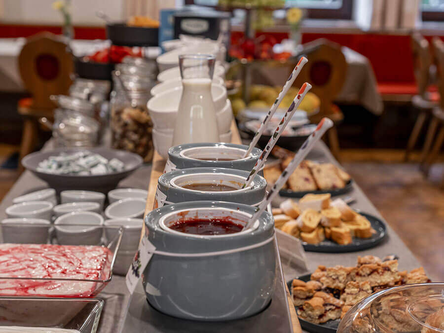 Ein Frühstücksbuffet mit Marmeladengläsern, Brot, Milch und Tellern, das auf einem langen Tisch in einem gemütlichen Speisesaal gedeckt ist. - Hotel Steinerhof