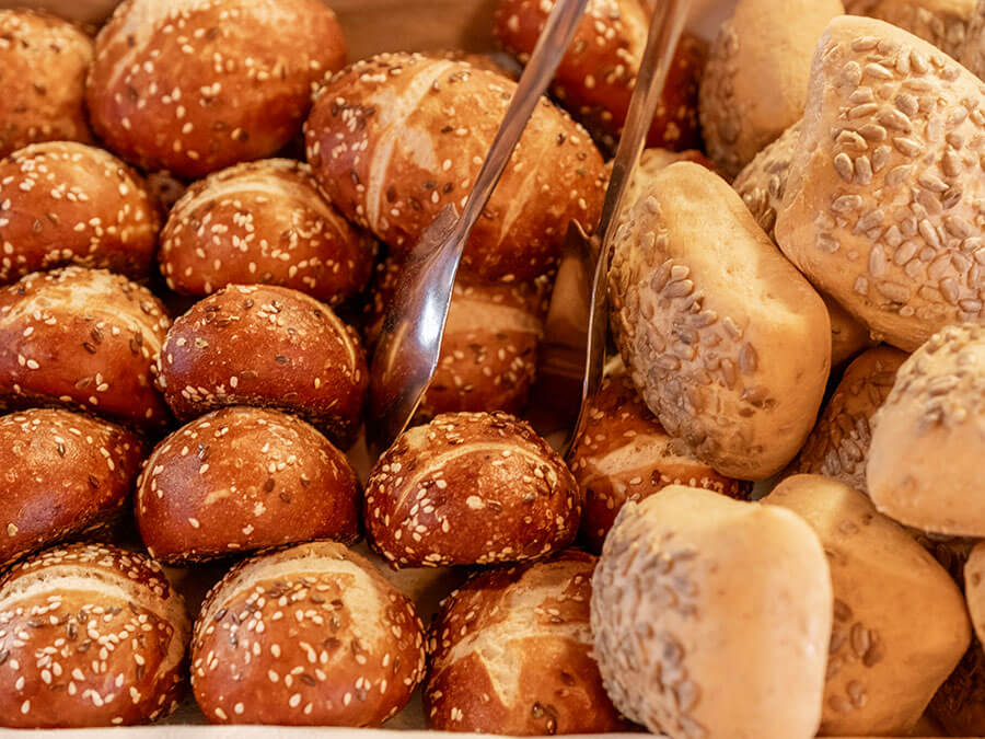 Verschiedene Brötchen mit Körnern, teilweise mit einer Zange, dicht nebeneinander in einer Bäckerei ausgestellt. - Hotel Steinerhof